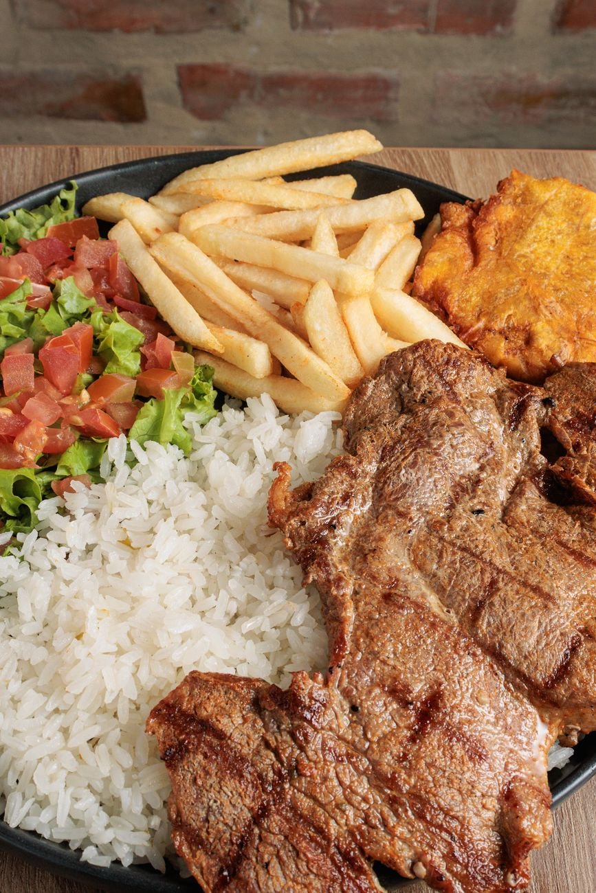 Colombian bandeja paisa with grilled meat, white rice, french fries, fried plantain, and fresh salad on a dark plate