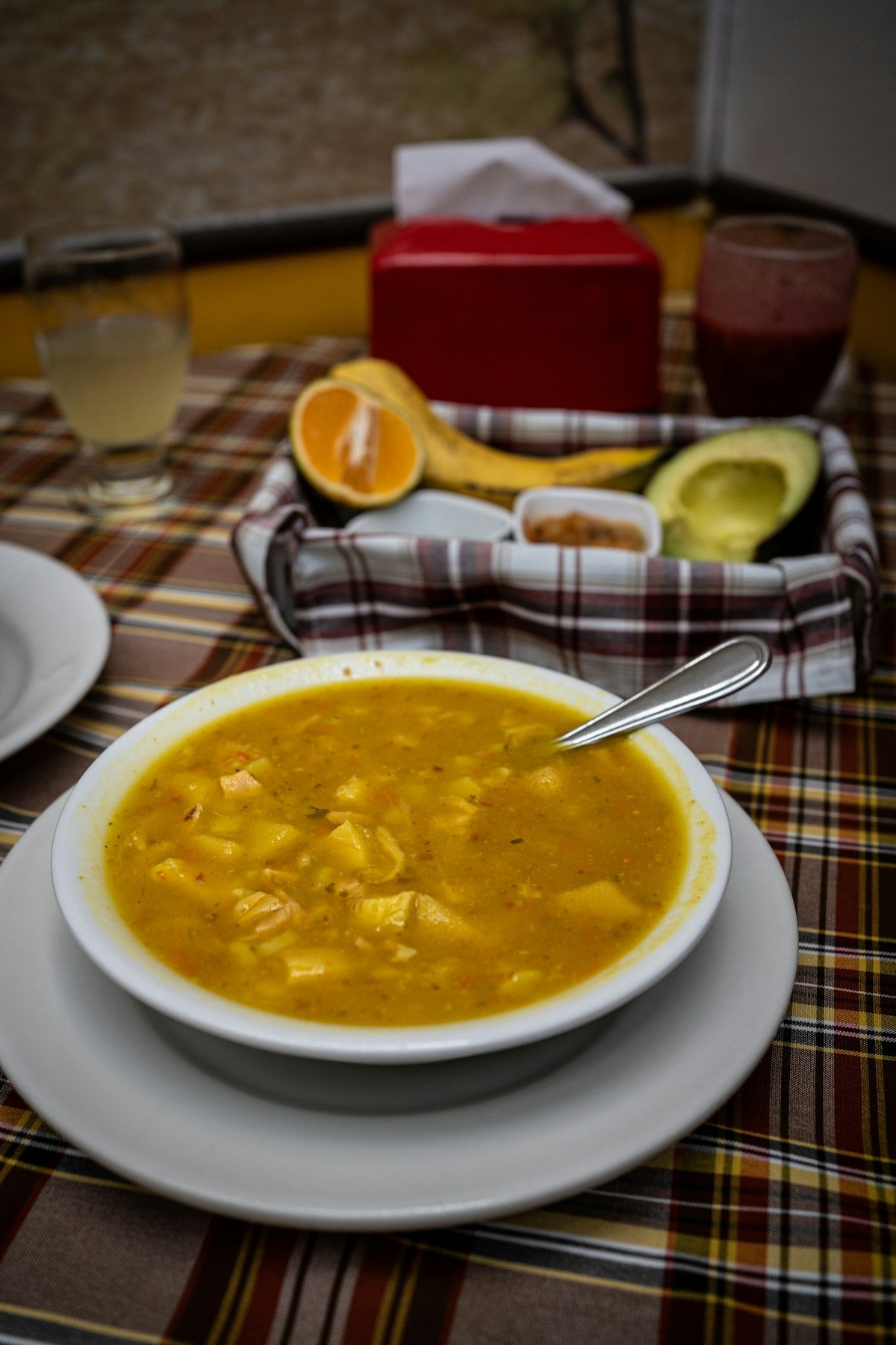 Golden sancocho soup with a spoon in a white bowl, with avocado, banana and citrus condiments on a plaid tablecloth, Colombia
