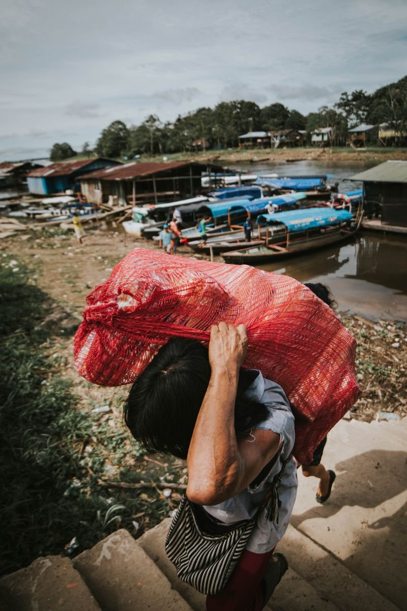 Person carrying goods at a busy Amazon river port with wooden boats moored along the muddy bank in the jungle
